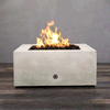A studio close-up showcases a square fire pit in a crisp white finish, resting on a dark wood floor. Bright orange flames rise continuously from a bed of black lava rocks, contrasting with the smooth texture of the concrete fire pit. A chrome key valve on the side panel indicates the manual control of this natural gas fire pit unit, demonstrating its clean design and functionality.