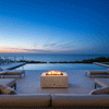 A luxury fire pit in a white finish sits centrally on a stone terrace, framed by a large sectional sofa and an infinity pool at twilight. The steady flames of the modern fire pit reflect on the water's surface, creating a sophisticated atmosphere for outdoor entertaining. The scene highlights the sleek design of this backyard oasis against a deep blue ocean horizon.