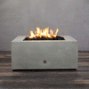 A close-up studio view presents a square fire pit cast in smooth grey concrete, resting on a dark wood floor against a matching wall. Bright yellow flames burn continuously from a bed of black lava rocks, showcasing the clean combustion and surface texture of the concrete fire pit. A chrome key valve is visible on the side panel, indicating the manual control mechanism of this natural gas fire pit unit.