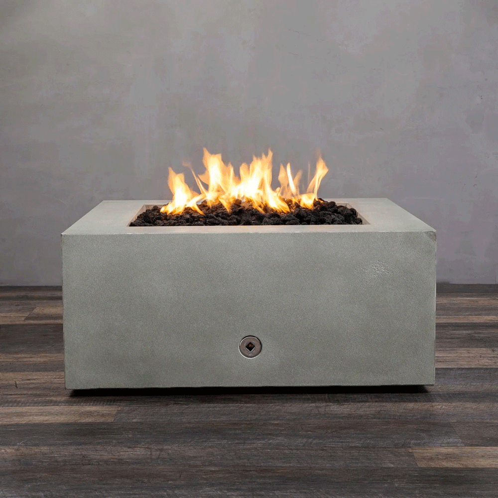 A close-up studio view presents a square fire pit cast in smooth grey concrete, resting on a dark wood floor against a matching wall. Bright yellow flames burn continuously from a bed of black lava rocks, showcasing the clean combustion and surface texture of the concrete fire pit. A chrome key valve is visible on the side panel, indicating the manual control mechanism of this natural gas fire pit unit.