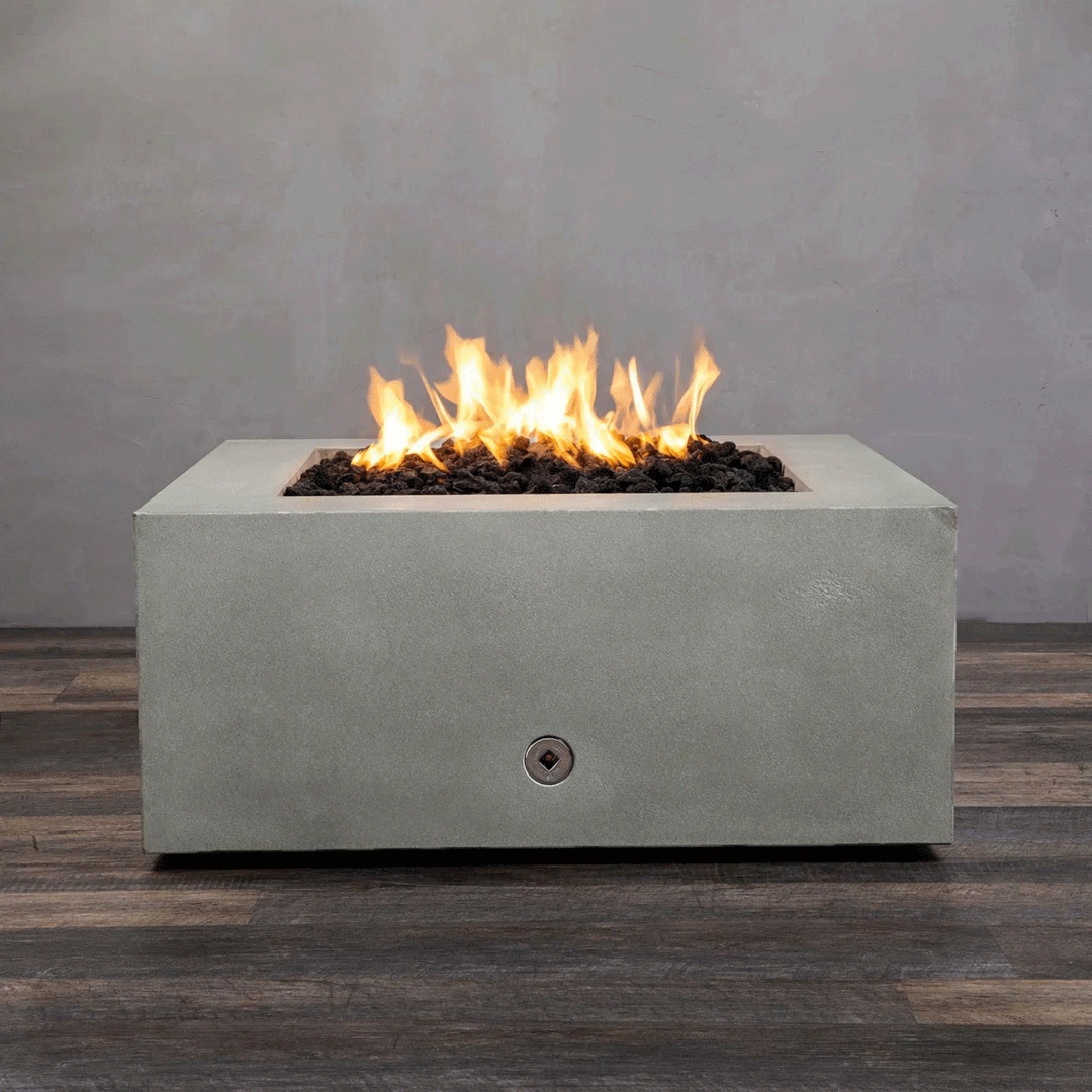 A close-up studio view presents a square fire pit cast in smooth grey concrete, resting on a dark wood floor against a matching wall. Bright yellow flames burn continuously from a bed of black lava rocks, showcasing the clean combustion and surface texture of the concrete fire pit. A chrome key valve is visible on the side panel, indicating the manual control mechanism of this natural gas fire pit unit.