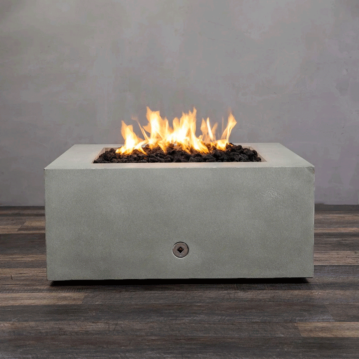 A close-up studio view presents a square fire pit cast in smooth grey concrete, resting on a dark wood floor against a matching wall. Bright yellow flames burn continuously from a bed of black lava rocks, showcasing the clean combustion and surface texture of the concrete fire pit. A chrome key valve is visible on the side panel, indicating the manual control mechanism of this natural gas fire pit unit.
