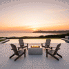 Four dark wood Adirondack chairs surround a white square fire pit on a patio overlooking the ocean and a distant lighthouse at sunset. The outdoor fire pit serves as a warm focal point, with bright flames rising from the center fire feature. This setup illustrates an idyllic family gathering fire pit location, where the gas fire pit enhances the scenic coastal view.