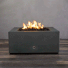 A close-up studio presentation displays a square fire pit cast in a matte graphite concrete, resting on a dark wood floor. Bright orange flames flicker continuously from a bed of black lava rocks, highlighting the smooth texture of the concrete fire pit. A chrome key valve on the side panel suggests a manual ignition system, demonstrating the clean design and functionality of this natural gas fire pit unit.