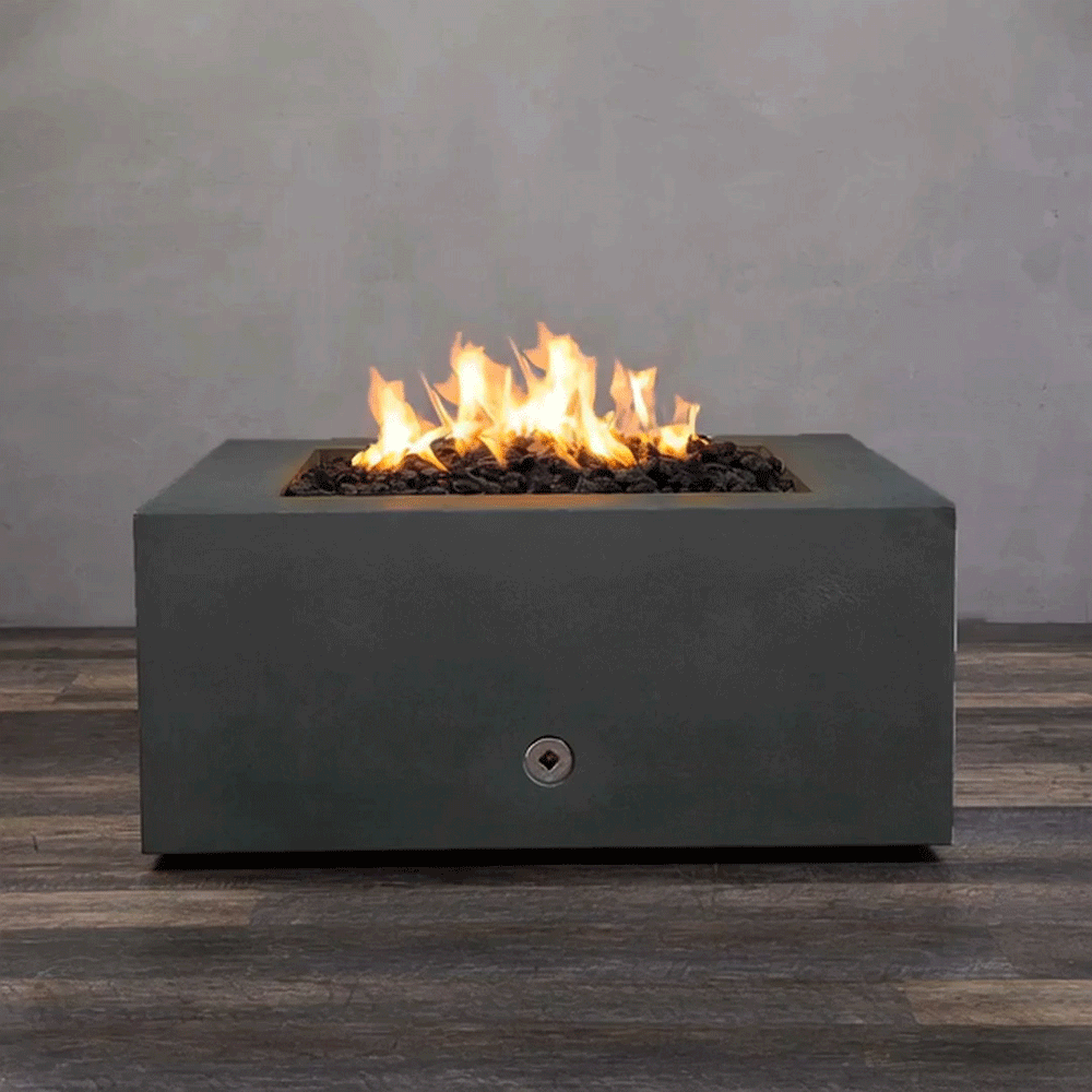 A close-up studio presentation displays a square fire pit cast in a matte graphite concrete, resting on a dark wood floor. Bright orange flames flicker continuously from a bed of black lava rocks, highlighting the smooth texture of the concrete fire pit. A chrome key valve on the side panel suggests a manual ignition system, demonstrating the clean design and functionality of this natural gas fire pit unit.