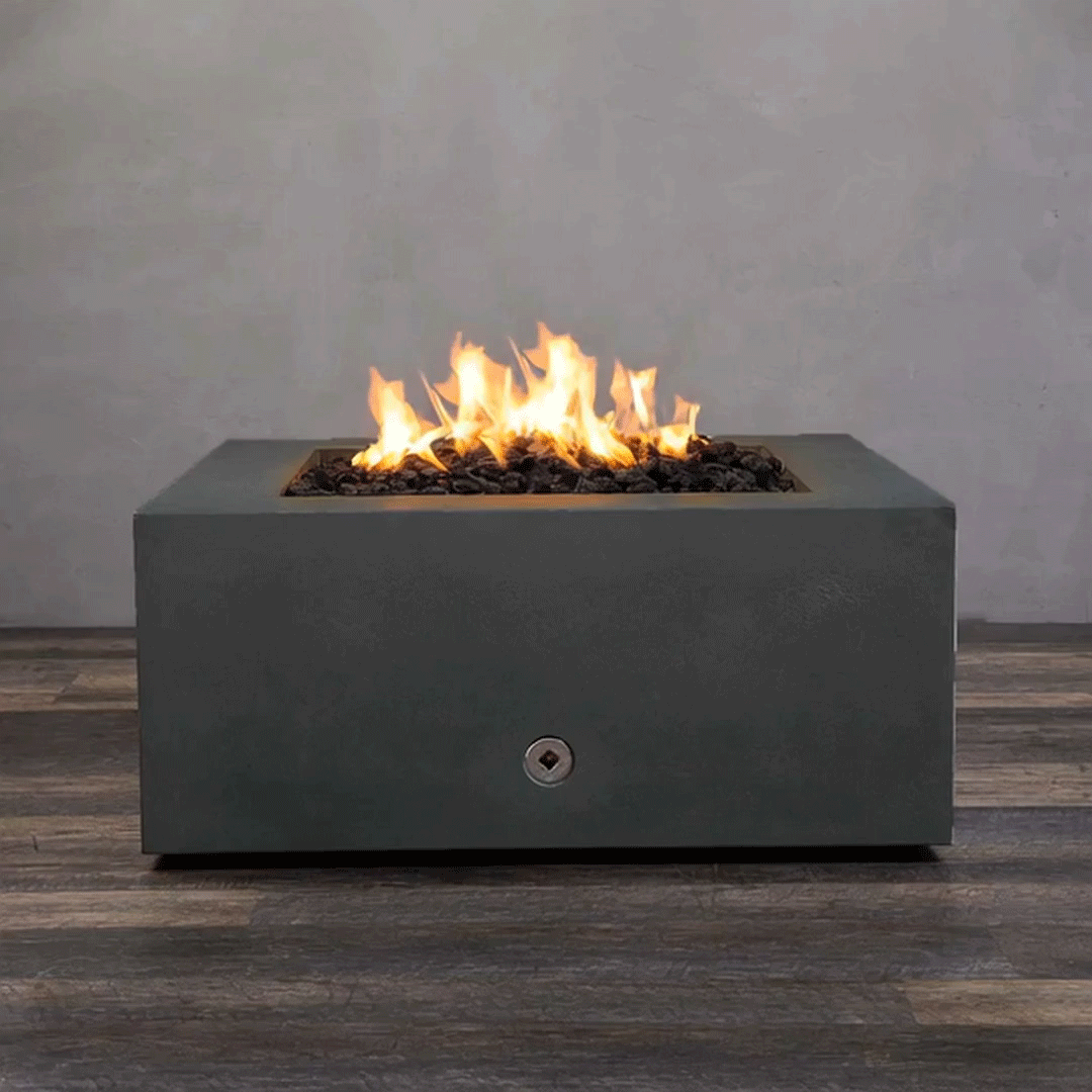A close-up studio presentation displays a square fire pit cast in a matte graphite concrete, resting on a dark wood floor. Bright orange flames flicker continuously from a bed of black lava rocks, highlighting the smooth texture of the concrete fire pit. A chrome key valve on the side panel suggests a manual ignition system, demonstrating the clean design and functionality of this natural gas fire pit unit.
