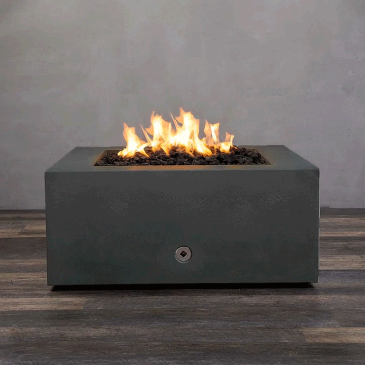 A close-up studio presentation displays a square fire pit cast in a matte graphite concrete, resting on a dark wood floor. Bright orange flames flicker continuously from a bed of black lava rocks, highlighting the smooth texture of the concrete fire pit. A chrome key valve on the side panel suggests a manual ignition system, demonstrating the clean design and functionality of this natural gas fire pit unit.