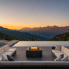 A U-shaped outdoor sectional sofa surrounds a low-profile square fire pit in a graphite finish, set against a spectacular mountain sunset. The patio fire pit is positioned on a paved deck next to an infinity pool, creating a luxurious backyard oasis. Clean-burning flames rise from the center, illustrating a smokeless fire pit experience that allows for unobstructed views and comfortable conversation in this spacious family gathering fire pit setting.