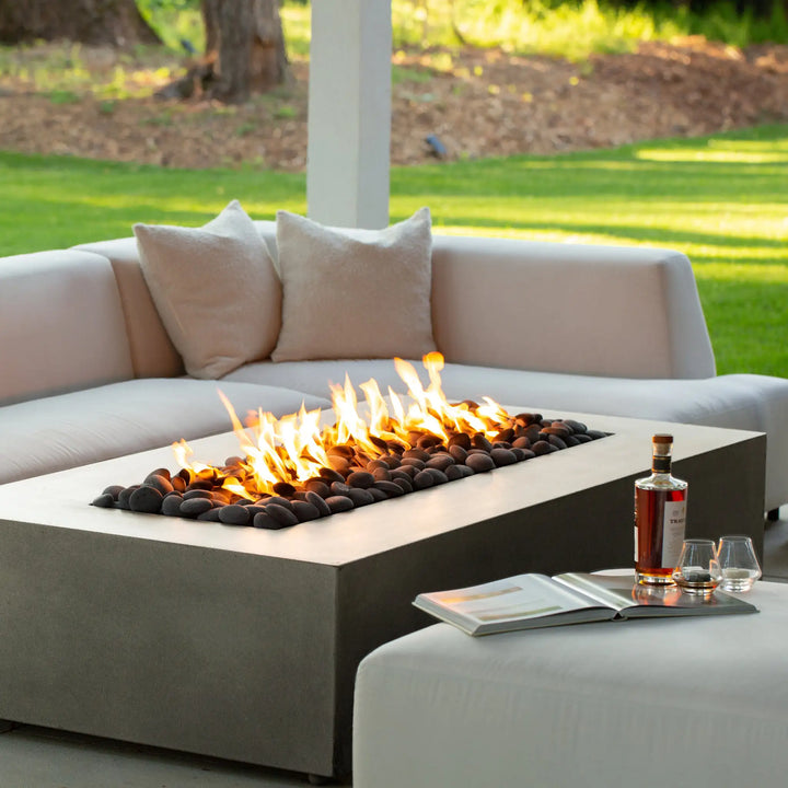 A serene lifestyle shot featuring the Starfire Designs Gravity 60" x 32" Rectangle Concrete Gas Fire Pit in beige, with a cozy seating arrangement and beverages set nearby. The flickering flames create a tranquil ambiance for relaxing or entertaining in outdoor spaces. This fire pit adds both style and function to any patio or backyard.