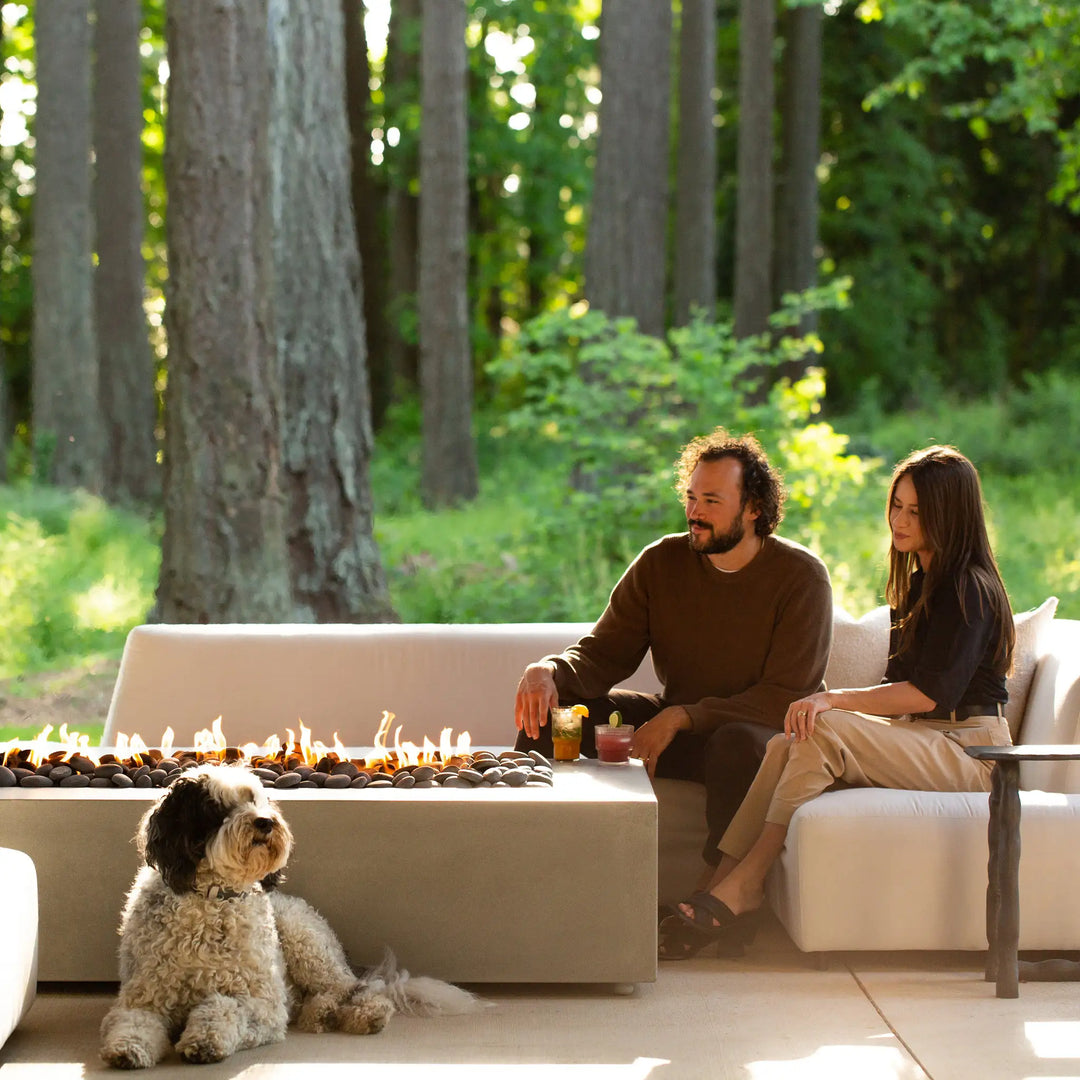 A lifestyle image of the Starfire Designs Gravity 60" x 32" Rectangle Concrete Gas Fire Pit in beige, set against a lush green forest backdrop. A couple enjoys drinks by the fire while a dog rests nearby, illustrating the fire pit's ability to bring warmth and ambiance to outdoor environments. Perfect for cozy evenings in nature.