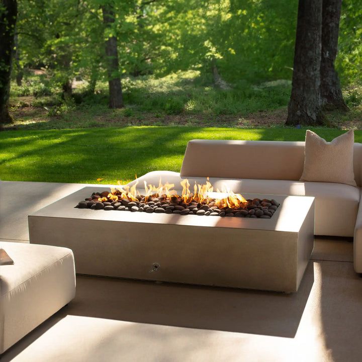 The Starfire Designs Gravity 60" x 32" Rectangle Concrete Gas Fire Pit in beige is displayed in an outdoor setting, with a beautiful flame glowing atop dark lava rocks. Set amidst plush outdoor furniture, the fire pit provides warmth and ambiance, making it the perfect centerpiece for social gatherings or quiet evenings outdoors.