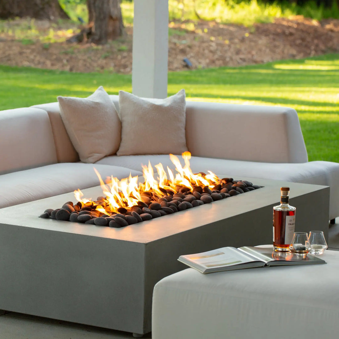 Stylish outdoor scene with the Starfire Designs Gravity 60" x 32" rectangle concrete gas fire pit in natural gray, set on a patio with beverages and books on a side table. The fire pit’s flames create a comfortable and relaxing atmosphere, perfect for intimate gatherings and evening entertaining. Its sleek design and premium materials make it an attractive focal point for any modern outdoor space.