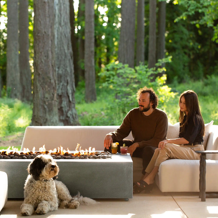 Forest setting with the Starfire Designs Gravity 60" x 32" rectangle concrete gas fire pit in natural gray. A cozy seating area with a dog lounging near the fire pit creates a relaxed, peaceful environment. The natural gray color of the fire pit complements the green surroundings, making it a great addition to outdoor spaces that blend nature with modern living.