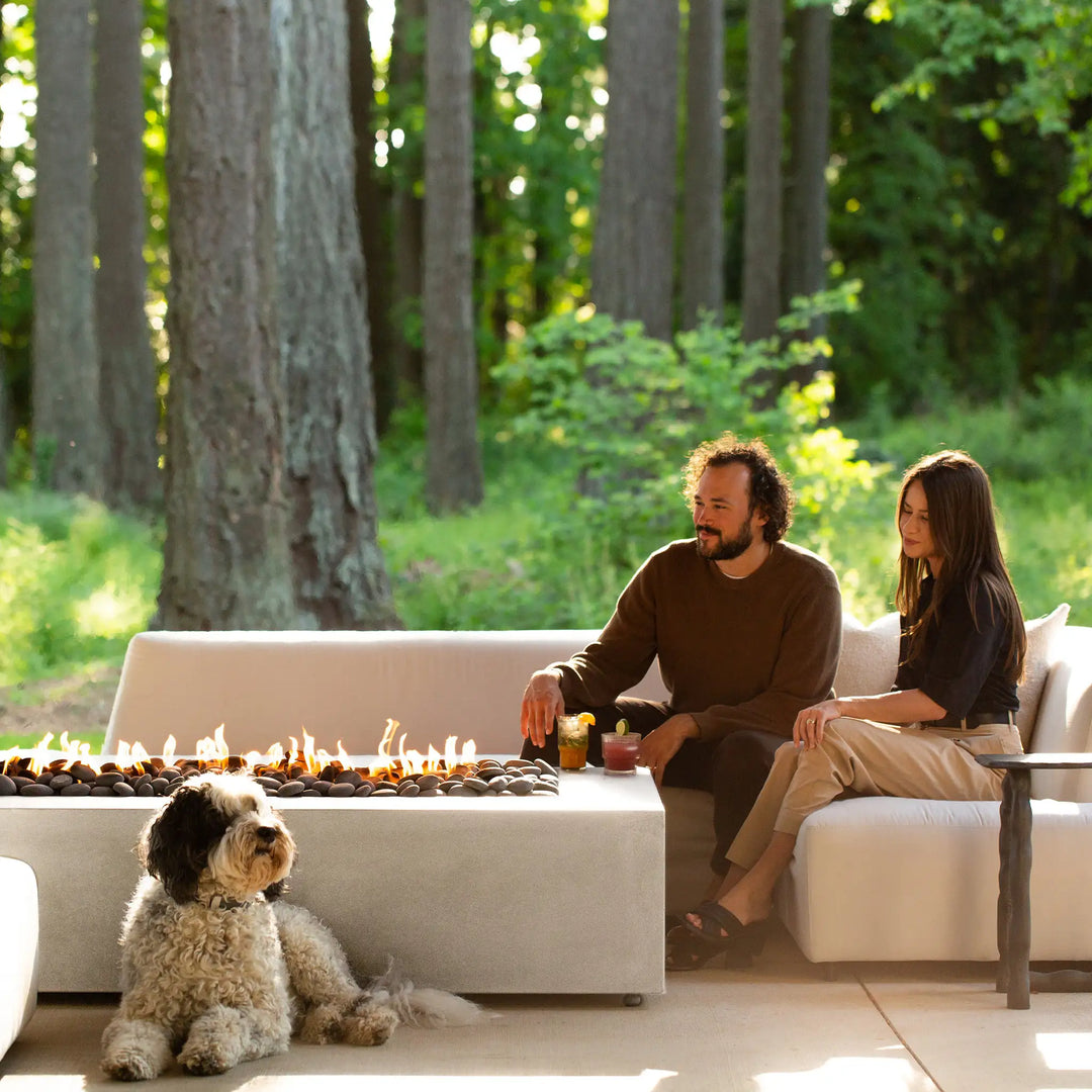 A lifestyle image of people relaxing by the Starfire Designs Gravity 60" x 32" Rectangle Concrete Gas Fire Pit in white, enjoying the warmth and ambiance. The fire pit enhances the modern outdoor seating arrangement, creating a perfect setting for social gatherings.