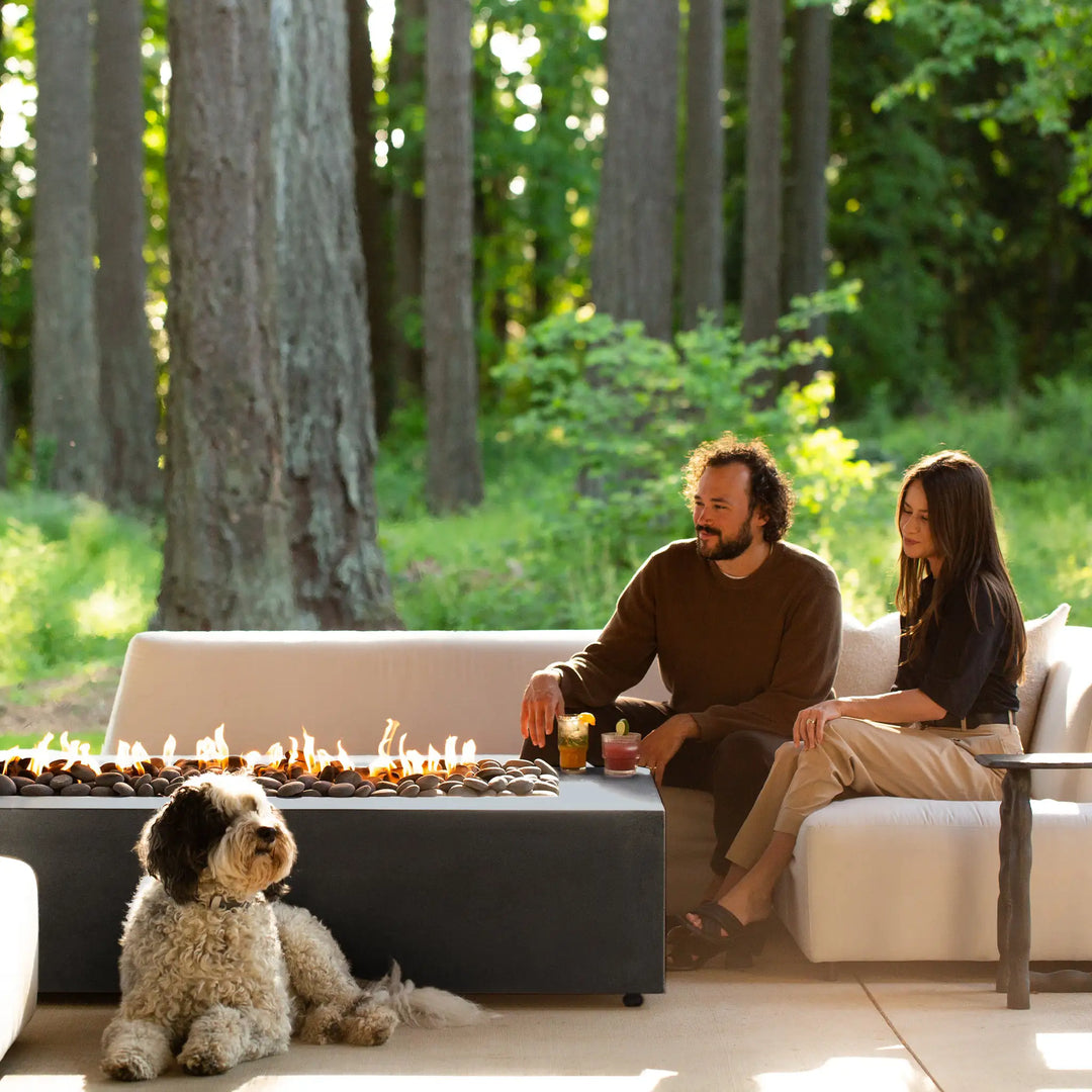 A serene outdoor scene with a family relaxing near the Starfire Designs Gravity 72-inch by 38-inch graphite concrete gas fire pit, set against a backdrop of lush greenery. The fire pit's vibrant flames and sleek graphite finish add warmth and ambiance to the outdoor setting, making it ideal for entertaining and relaxing in style.