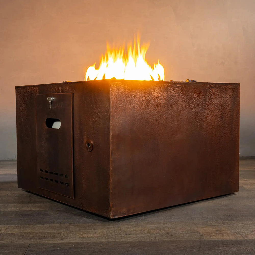 Side view of the 36-inch Gravity Moreno square copper fire pit by Starfire Designs, showcasing its hammered copper texture and vibrant flames in a cozy indoor setting