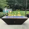 A luxurious outdoor fire pit table filled with Starfire Glass 1/2" Broken Fire Glass (10lbs) in Pacific Blue Reflective set against a lush backyard. The brilliant reflective fire glass intensifies the warm flames, creating a relaxing ambiance for outdoor gatherings. Perfect for propane fire tables, backyard fire pits, and modern patio fire bowls.