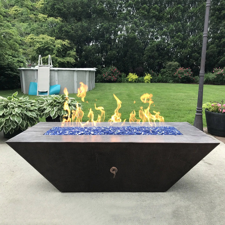 A luxurious outdoor fire pit table filled with Starfire Glass 1/2" Broken Fire Glass (10lbs) in Pacific Blue Reflective set against a lush backyard. The brilliant reflective fire glass intensifies the warm flames, creating a relaxing ambiance for outdoor gatherings. Perfect for propane fire tables, backyard fire pits, and modern patio fire bowls.