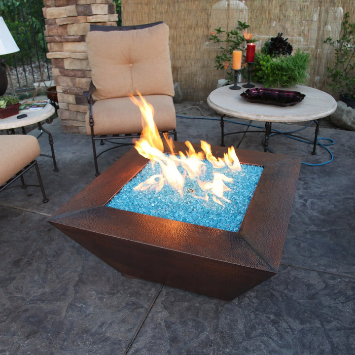 A beautifully designed square fire pit table filled with Starfire Glass 1/2" Broken Fire Glass (10lbs) in Pacific Blue Reflective, glowing under a cozy outdoor patio setup. The high-end reflective glass maximizes flame brilliance, making it a must-have for luxury fire pits, outdoor patio heating, and modern fire features.