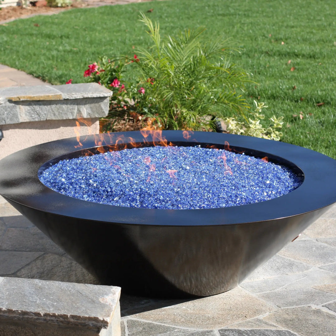 A sleek black outdoor fire bowl filled with shimmering blue Starfire Glass 1/4" broken fire glass (10lbs), situated on a patio surrounded by lush greenery. Perfect for modern outdoor settings. 