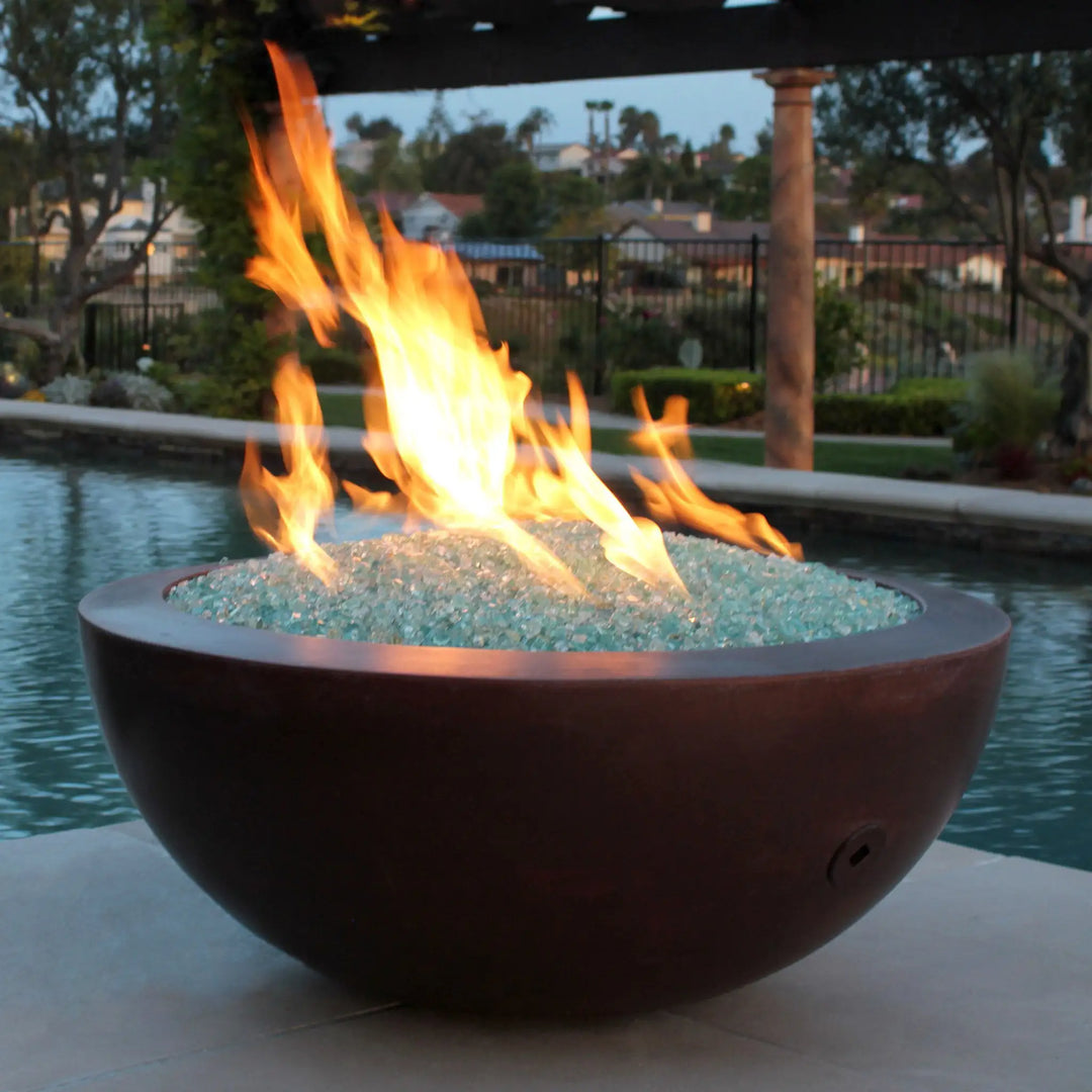 A curved copper fire bowl filled with reflective aqua Starfire Glass 1/4" broken fire glass (10lbs), glowing warmly by a serene poolside. A picturesque setting for outdoor gatherings. 
