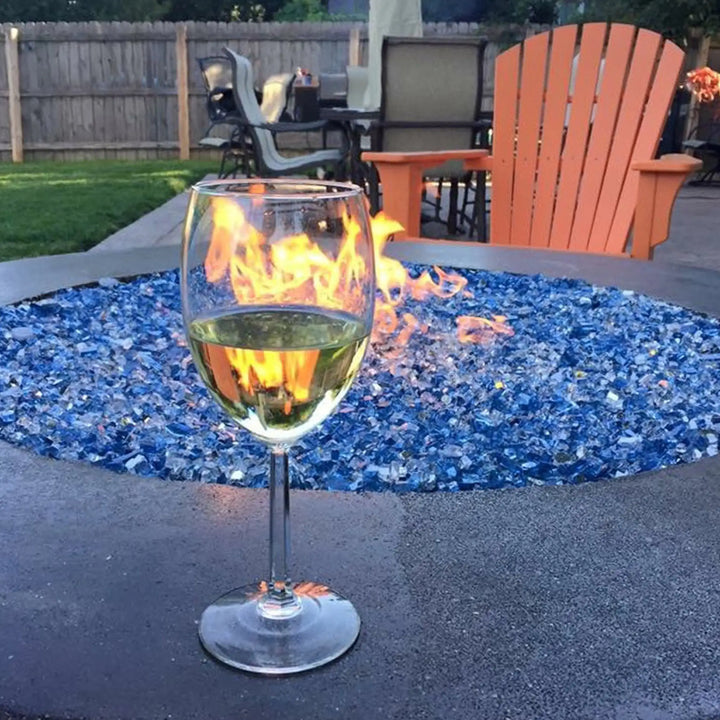 A vibrant backyard patio featuring a round fire pit filled with sparkling blue Starfire Glass 1/4" broken fire glass (10lbs). A glass of white wine rests on the fire pit edge, creating a cozy and inviting outdoor ambiance.