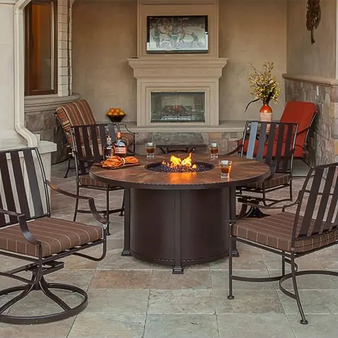 Outdoor seating area with a round fire pit table surrounded by four metal-framed chairs with brown cushions, set on a covered patio with a stone fireplace in the background and drinks and snacks on the table