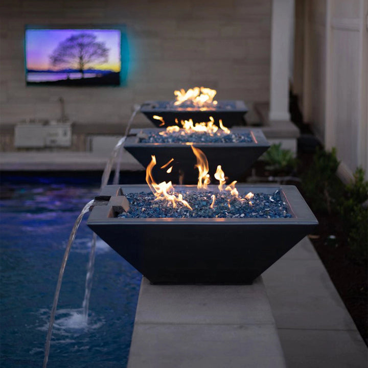 A stunning outdoor setup featuring multiple The Outdoor Plus Maya Square Concrete Fire and Water Bowls in slate finish. Each bowl is elegantly positioned along a poolside, showcasing vibrant blue fire glass and water flowing from the bowls into the pool below. The scene is illuminated by the warm glow of fire against a modern backdrop, creating a serene and luxurious outdoor atmosphere perfect for entertaining or relaxation.
