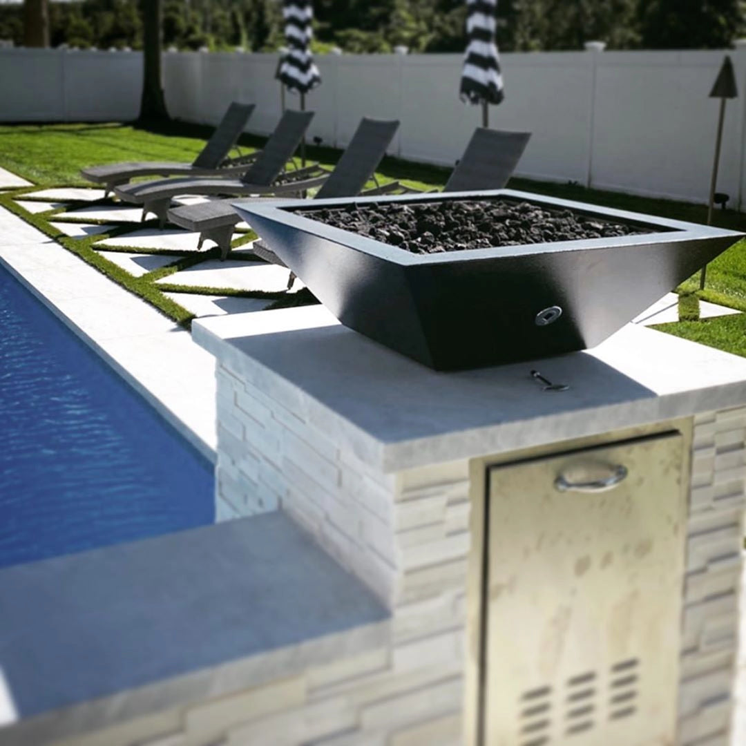 The Outdoor Plus 24-inch Square Concrete Maya Fire Bowl in a sleek black finish, placed on a poolside stone pillar with a modern outdoor landscape. Featuring dark lava rocks and a smooth, minimalist design, this fire bowl adds a contemporary touch to the outdoor seating area, perfect for enhancing poolside ambiance