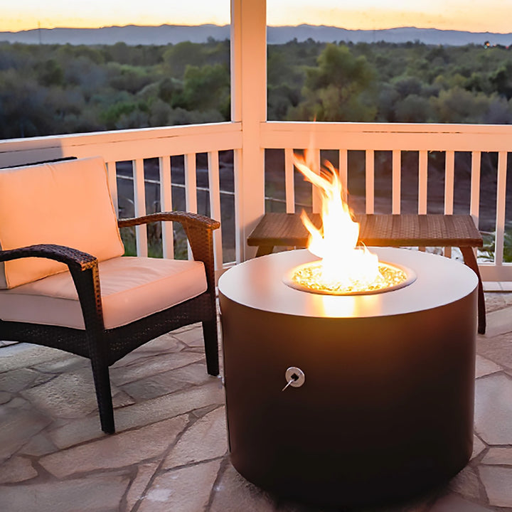 A stunning view of the Outdoor Plus 30" Beverly Steel Gas Fire Pit on a deck at sunset, offering warmth and elegance. The fire pit, in a sleek dark finish, stands out against the backdrop of nature, providing the perfect setting for a relaxing evening outdoors. Comfortable seating surrounds the fire pit for cozy outdoor lounging.