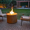 A serene evening garden setting with the Outdoor Plus 30" Beverly Steel Gas Fire Pit in corten steel finish, radiating a warm glow. The fire pit is set in the center of a brick-paved patio, providing a perfect ambiance for a cozy outdoor gathering. The surrounding greenery and wooden pergola complete the peaceful atmosphere.