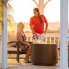 Two people enjoying the warmth of the Outdoor Plus 30" Beverly Steel Gas Fire Pit at sunset in a gazebo. The fire pit, in a powder-coated steel finish, sits on a stone floor, offering a relaxing setting. The image captures a peaceful moment with the fire pit creating a focal point for the gathering.