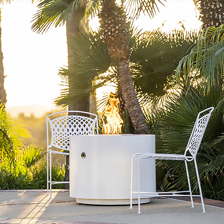 The Outdoor Plus 30" Beverly Steel Gas Fire Pit in a white powder-coated finish, set against a backdrop of palm trees and bright sunshine. The fire pit adds a modern touch to this tropical outdoor setting, providing a focal point for enjoying the sunny environment. Metal chairs surround the fire pit for casual seating.