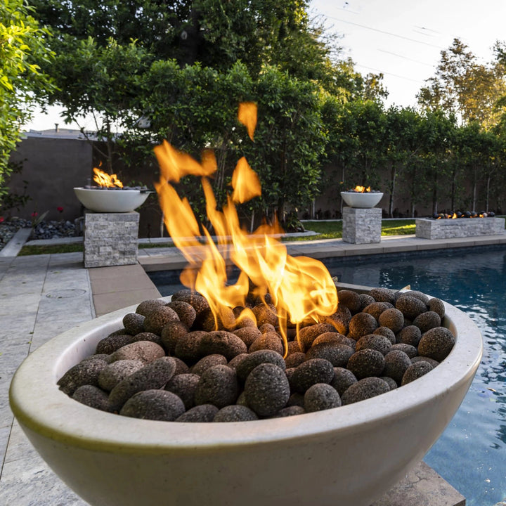 A close-up view of The Outdoor Plus 30" Luna Concrete Gas Fire Pit, showcasing its use by a modern poolside setup. The fire pit is filled with natural lava rocks, with a large flame rising, creating a warm ambiance for outdoor relaxation. Its sleek, round concrete design complements the minimalist backyard landscaping. Perfect for patios, poolsides, or garden spaces, this gas fire pit adds both warmth and a stylish touch to any outdoor setting