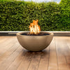 A modern The Outdoor Plus 30" Luna Concrete Gas Fire Pit displayed on a wooden deck with greenery in the background. This round gas fire pit features a large flame set atop black lava rocks, providing an inviting warmth for outdoor gatherings. Its durable concrete finish is perfect for contemporary outdoor designs, adding a cozy and functional focal point to any patio or garden area. Ideal for entertaining, this fire pit creates an elegant and warm atmosphere.