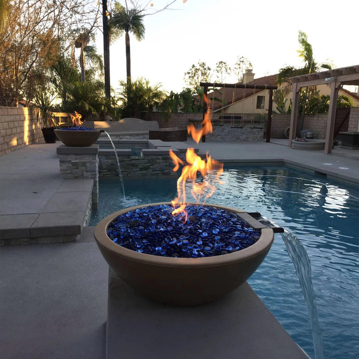 A luxurious Outdoor Plus 33" Round Concrete Sedona Fire and Water Bowl in a sleek black finish installed beside a pool. The fire bowl features a built-in waterfall spout, creating a stunning visual effect as water flows into the pool, while a bright flame burns above. Perfect for modern backyard designs, this fire and water bowl is an elegant centerpiece. Ideal for use with natural gas or propane, providing both fire and water elements for outdoor ambiance