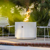 A stylish poolside setting with The Outdoor Plus 36" Beverly Steel Gas Fire Pit in a white powder-coated finish. The fire pit stands elegantly on the patio, paired with minimalist white outdoor chairs, providing warmth and a modern aesthetic to the space. Its sleek round shape makes it an ideal focal point for poolside relaxation.