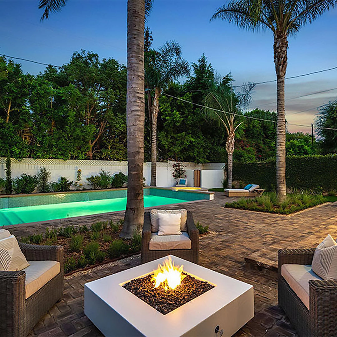 The Outdoor Plus 36-inch Cabo Square Concrete Gas Fire Pit is featured in this lifestyle image with a white finish, located poolside amidst palm trees and lush greenery. The fire pit is in full operation, with soft glowing flames adding elegance and warmth to the outdoor setting. Surrounded by cozy wicker chairs, this fire pit enhances the backyard ambiance, making it perfect for outdoor gatherings and relaxing evenings. The fire pit's sleek white surface and durable construction are ideal for high-end