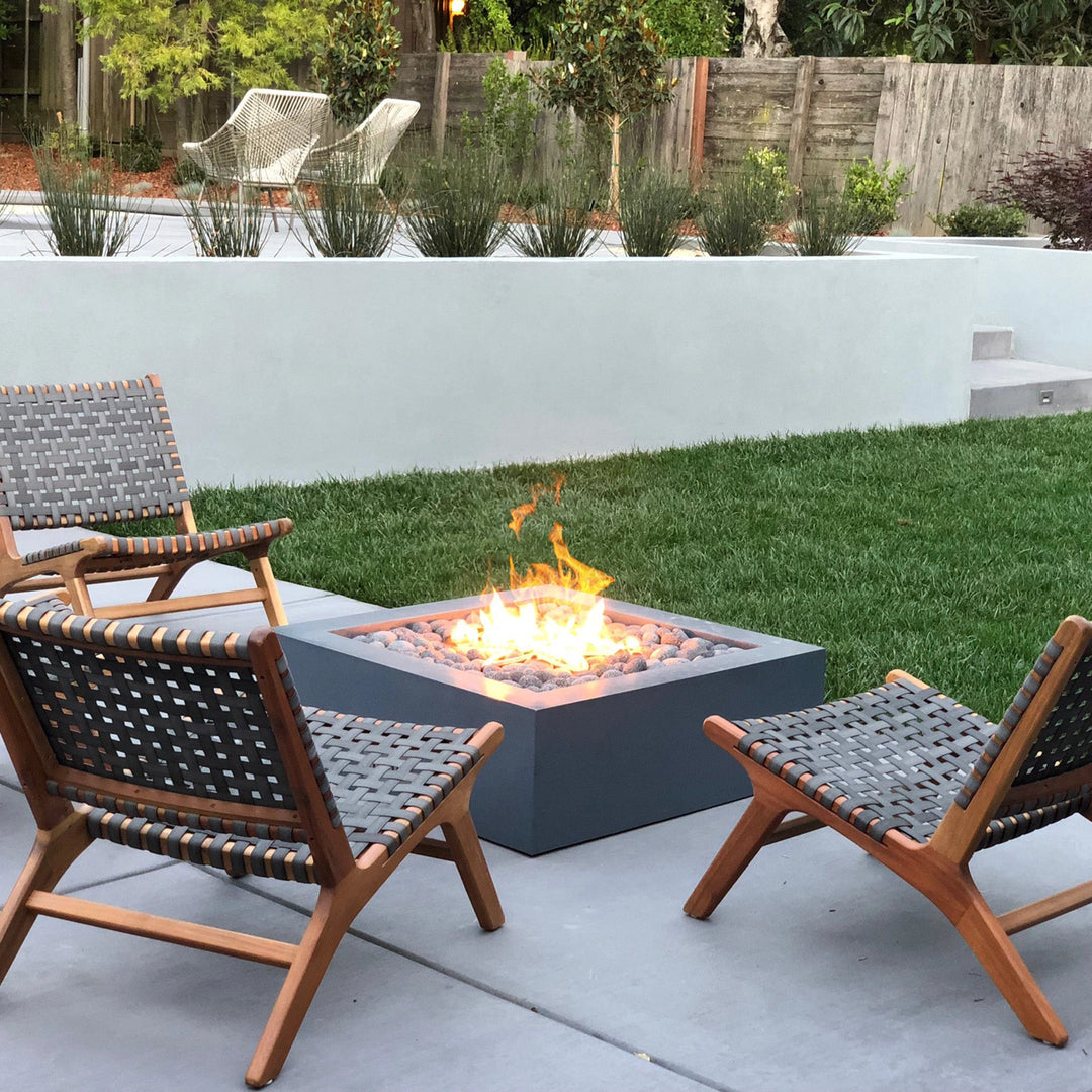 A modern backyard setting featuring the Outdoor Plus 36" Quad Concrete Gas Fire Pit with a clean geometric design. This square gas fire pit is surrounded by minimalist patio furniture, with wooden chairs and woven accents, providing a cozy ambiance. The fire pit is lit, showcasing a natural gas flame with stone pebbles. Perfect for outdoor fire pit enthusiasts looking to enhance their backyard or patio with a modern touch. Ideal for luxury outdoor decor, patio heating, and backyard design.