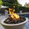 The Outdoor Plus 38-inch Luna concrete gas fire pit placed poolside, surrounded by a luxurious outdoor setting with multiple fire features in view. The large smooth stones inside the bowl emphasize its contemporary design, while vibrant flames provide an eye-catching focal point. Ideal for high-end outdoor living spaces, this concrete gas fire pit enhances the ambiance of patios, pool areas, and gardens.