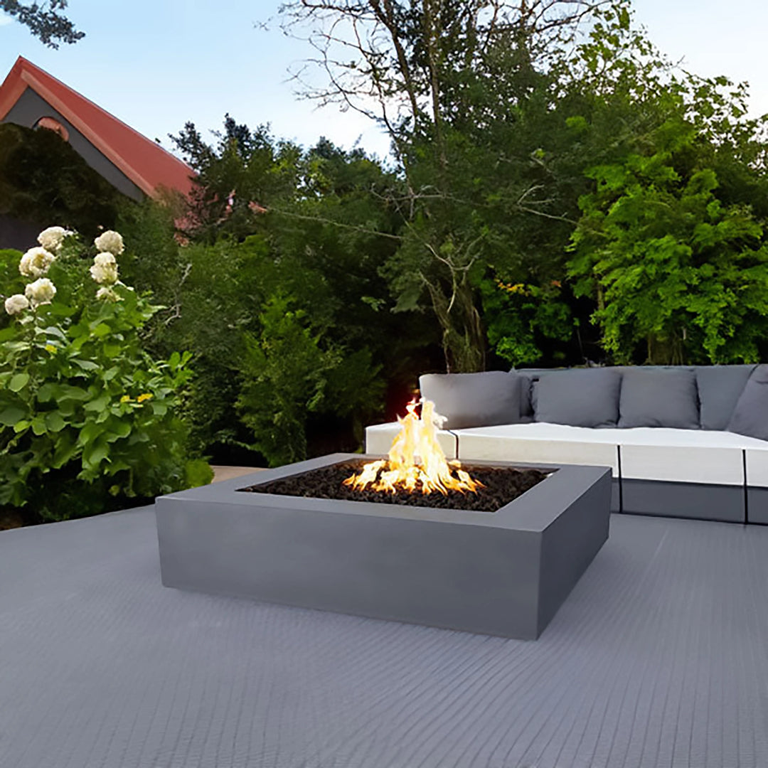 This image features The Outdoor Plus 42" Quad Concrete Gas Fire Pit placed in a stylish backyard setting. The modern fire pit, with its clean, square design, showcases the gray concrete finish that blends well with the outdoor couch seating. The fire pit's vibrant flame creates a cozy and inviting atmosphere, perfect for relaxing and entertaining.