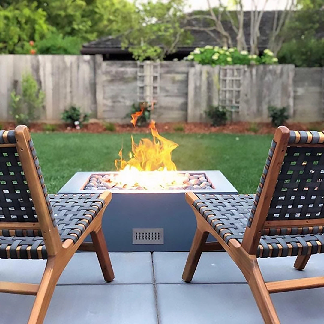 The Outdoor Plus 42" Quad Concrete Gas Fire Pit is showcased in a mid-century modern patio setting. Surrounded by woven lounge chairs, the fire pit takes center stage with its sleek square design and smooth gray finish. The burning fire adds warmth and ambiance to the outdoor environment, making it ideal for evening gatherings.