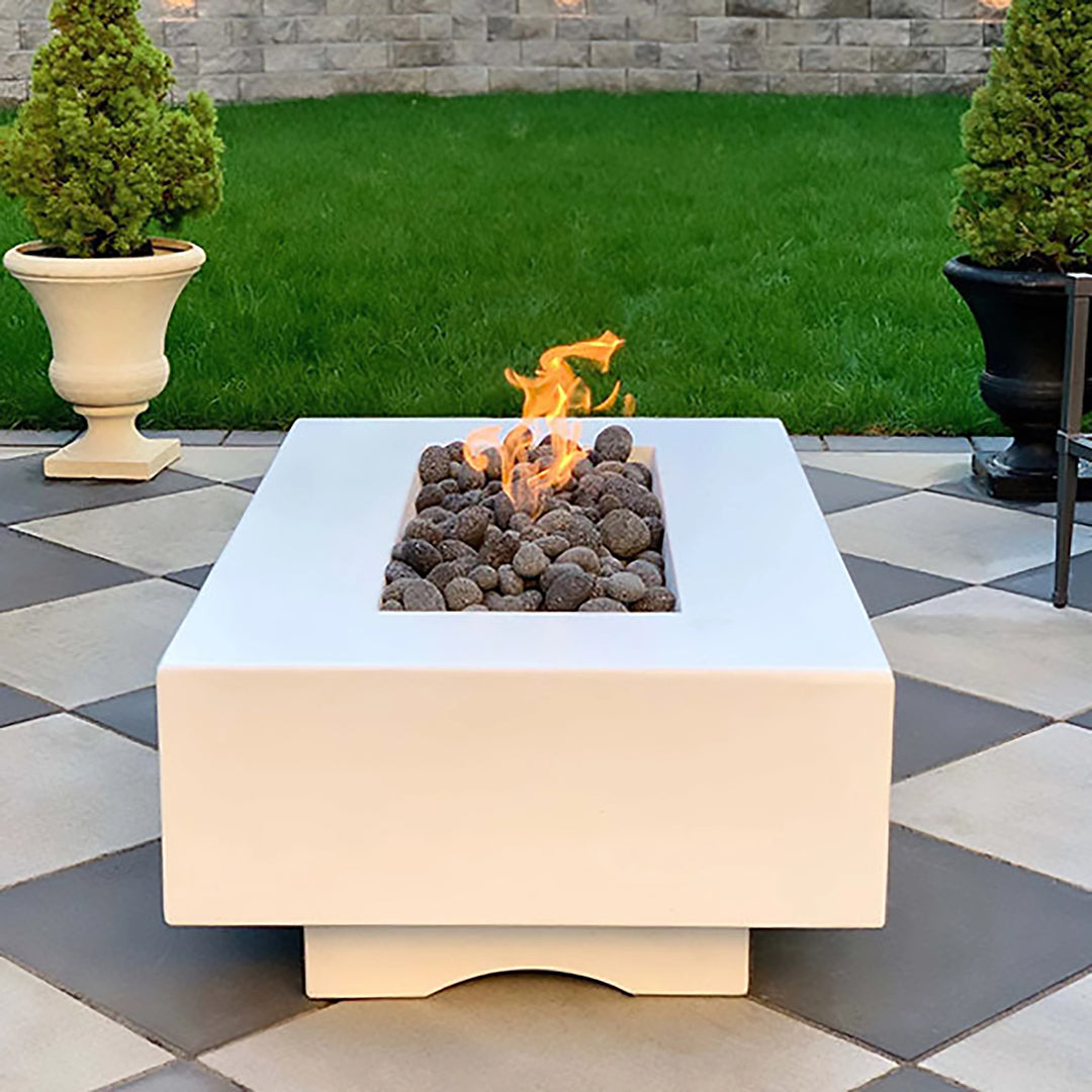 The Outdoor Plus 48" Del Mar Concrete Gas Fire Pit in white, positioned as the centerpiece in a landscaped backyard. The modern fire pit design is accentuated by the lush green grass and carefully manicured garden. The flickering flames create a cozy atmosphere, perfect for outdoor gatherings and luxury backyard setups. Suitable for modern fire pit tables, patio fire features, and sophisticated outdoor decor.