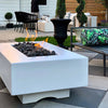 Close-up shot of The Outdoor Plus 48" Del Mar Concrete Gas Fire Pit in white, positioned in a backyard setup with flames dancing over smooth black lava rocks. The rectangular shape and clean lines of the fire pit contrast with the surrounding natural elements. Ideal for modern fire pit designs, outdoor fire pit tables, and creating a warm ambiance in upscale backyard spaces