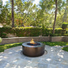 The Outdoor Plus 48" Round Unity Fire Pit shown in a modern garden setting. This outdoor fire pit features a sleek round design with a matte black finish, blending seamlessly into the natural surroundings. Perfect for enhancing your outdoor space with warmth and ambiance, this luxury fire pit is ideal for contemporary patio setups. The Unity fire pit is a versatile addition to any backyard or landscape, providing a central gathering point for family and friends.