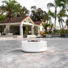 The Outdoor Plus 48" Round Unity Fire Pit in a white concrete finish, accentuating a luxurious patio with a water feature in the background. This high-end fire pit offers a blend of elegance and utility, making it ideal for outdoor spaces designed for relaxation and entertainment. The Unity fire pit's robust construction and refined appearance ensure it stands out as a focal point, providing a welcoming warmth for any outdoor living area. 