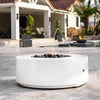 The Outdoor Plus 48" Round Unity Fire Pit in a white concrete finish, displayed on a spacious modern patio with a fountain feature in the background. This contemporary fire pit is crafted from durable materials, offering both aesthetic appeal and functionality for outdoor gatherings. The clean, minimalist design of this fire pit complements modern architecture, making it a perfect centerpiece for upscale homes.