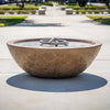 The Outdoor Plus 48-inch Sedona Concrete Gas Fire Pit, showcased in a backyard setting with lush greenery and colorful flowers. The bowl-shaped fire pit is mounted on a stone pedestal, and its smooth finish in a dark color contrasts beautifully with the natural stone base. Filled with decorative pebbles and equipped with a flame, this fire pit creates a warm and inviting atmosphere. Ideal for patios, backyard fire features, and enhancing the ambiance of outdoor gatherings with its modern fire pit design
