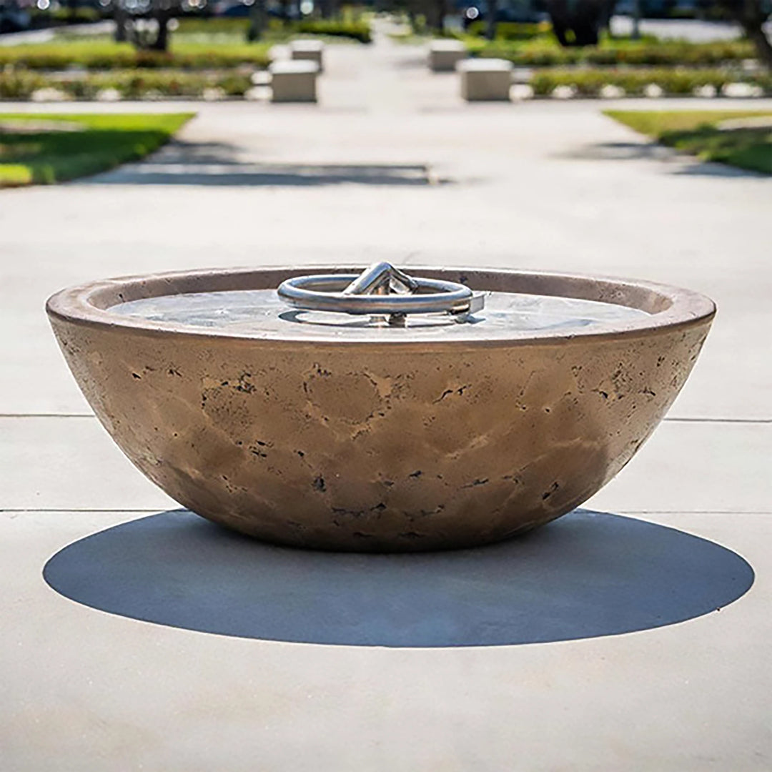The Outdoor Plus 48-inch Sedona Concrete Gas Fire Pit, showcased in a backyard setting with lush greenery and colorful flowers. The bowl-shaped fire pit is mounted on a stone pedestal, and its smooth finish in a dark color contrasts beautifully with the natural stone base. Filled with decorative pebbles and equipped with a flame, this fire pit creates a warm and inviting atmosphere. Ideal for patios, backyard fire features, and enhancing the ambiance of outdoor gatherings with its modern fire pit design