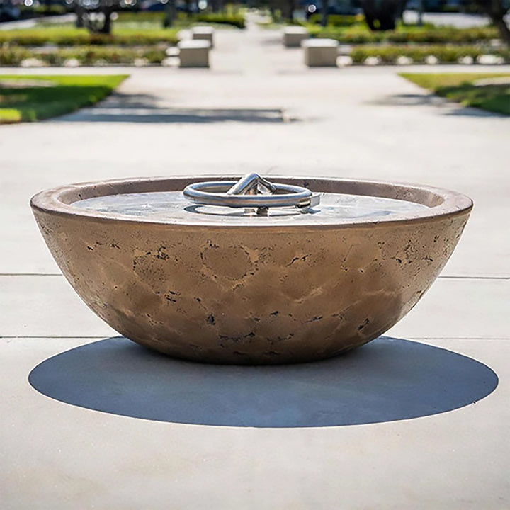The Outdoor Plus 48-inch Sedona Concrete Gas Fire Pit, showcased in a backyard setting with lush greenery and colorful flowers. The bowl-shaped fire pit is mounted on a stone pedestal, and its smooth finish in a dark color contrasts beautifully with the natural stone base. Filled with decorative pebbles and equipped with a flame, this fire pit creates a warm and inviting atmosphere. Ideal for patios, backyard fire features, and enhancing the ambiance of outdoor gatherings with its modern fire pit design